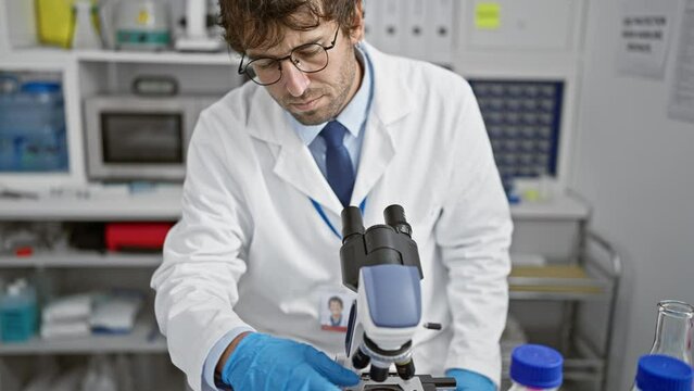 Serious young, handsome hispanic man scientist examining and analysing a medical sample at his clinic's lab using a microscope, in a quest for groundbreaking discovery.