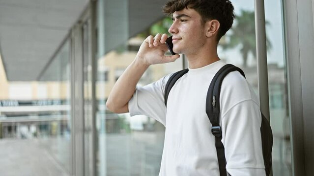 Casual-cool young hispanic university student, using smartphone for an important call with his backpack on, standing in sunlit urban campus, immersed in conversation.