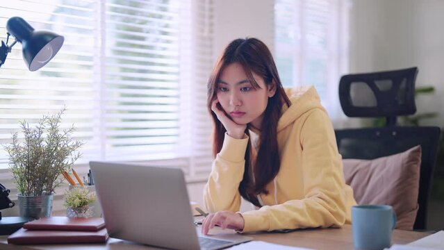 Asian female student reads study materials on her laptop with a hint of boredom at her home desk. Capture relatable moments of home study for educational projects.