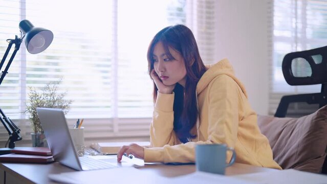 Asian female student reads study materials on her laptop with a hint of boredom at her home desk. Capture relatable moments of home study for educational projects.