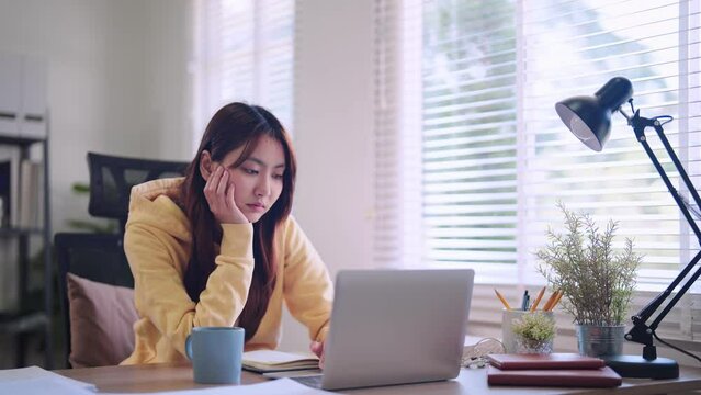 Asian female student engaged in online learning on her laptop at the home desk, battling drowsiness and eventually dozing off