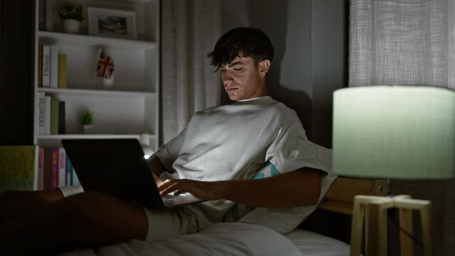 Handsome young hispanic teenager comfortably resting, concentrated on laptop, engulfed in the morning light, sitting relaxed on his cozy bed in a cool room.