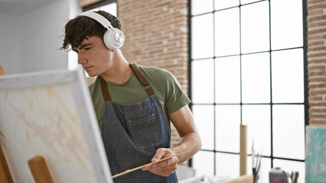 Handsome young hispanic art student immersed in his creativity, fervently drawing at an indoor studio, expertly mastering paintbrush and palette while listening to inspirational music.