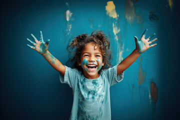 A curly-haired girl laughs directly at the camera and shows her hands stained with multi-colored paints on a colored studio background. Happy childhood concept.