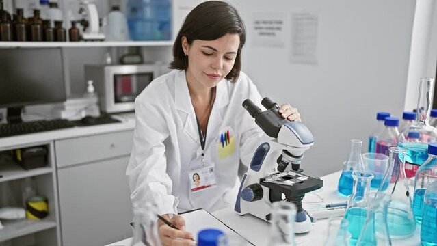 Energetic young hispanic woman scientist with a beautiful hairstyle, engrossed in her medical research, meticulously taking notes while peering through a microscope at a bustling laboratory.