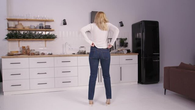 Rear view of disappointed pretty middle aged woman in high heels trying to squeeze into old tight jeans after gaining weight, expressing frustration and despair while standing in domestic kitchen.