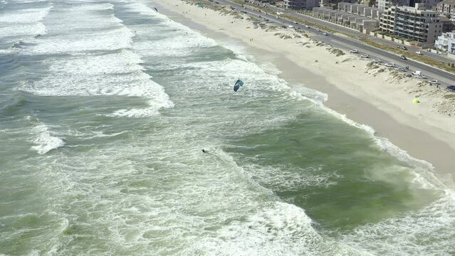 Drone of beach, people kite surfing on waves and city background for travel, vacation and summer holiday. Aerial view of sea water, surfer in ocean and cars driving on road in Cape Town, South Africa