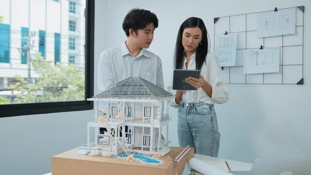 Beautiful young caucasian project manager brief asian engineer by using tablet while engineer measuring house model on meeting table with blueprint, architectural equipment scatter around. Immaculate