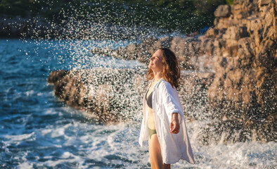 Young cheerful relaxed woman in a white shirt enjoying the sea waves and splashes while standing on shore