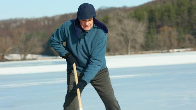 An elderly man practices stricking the puck with hockey sticks on a frozen lake in winter.
