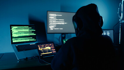 A hacker breaks into company data servers in dark atmosphere and has multiple displays. Hacker in dark room surrounded computers. Hacker breaks into corporate data server. Digital system security