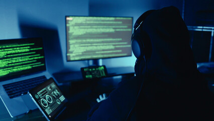 A hacker breaks into company data servers in dark atmosphere and has multiple displays. Hacker in dark room surrounded computers. Hacker breaks into corporate data server. Digital system security