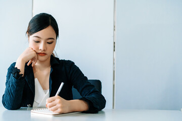 Beautiful young Asian woman writes letter on notebook while sitting at office desk. Content writer and secretary job concept. uds - obrazy, fototapety, plakaty