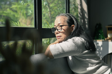 Pensive senior woman sitting on couch and looking in distance out of window.