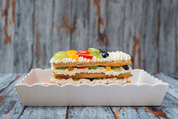 Mille-feuille cake decorated with fruit served on a white platter. Wooden background.