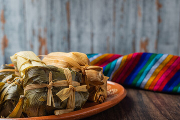 Different tamales in a clay dish on wooden table with a colorful serape.
