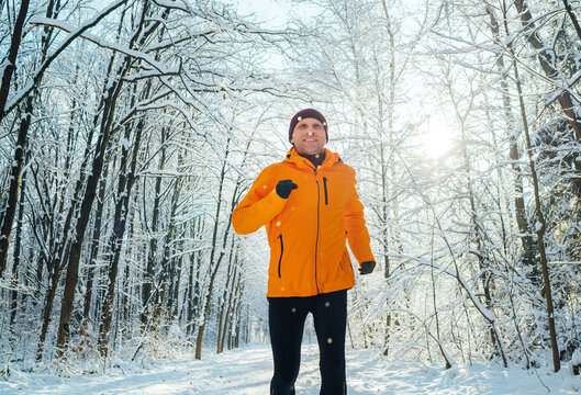 Middle-aged smiling trail runner man dressed bright orange windproof jacket endurance running picturesque snowy forest. Sporty active people and winter training concept image.