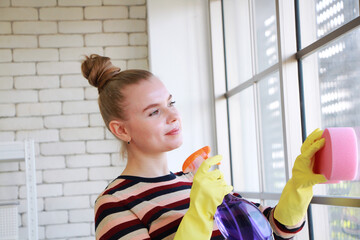 Smiling Young Caucasian woman Wear yellow rubber gloves is holding Cleanser bottle and sponge. She is cleaning and wiping the glass window for housework. Housewife lifestyle on holiday concept - Powered by Adobe