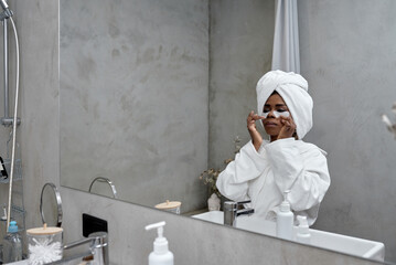Attractive afro woman wears hydrogel patches under eyes dressed in bathrobe and wrapped towel....