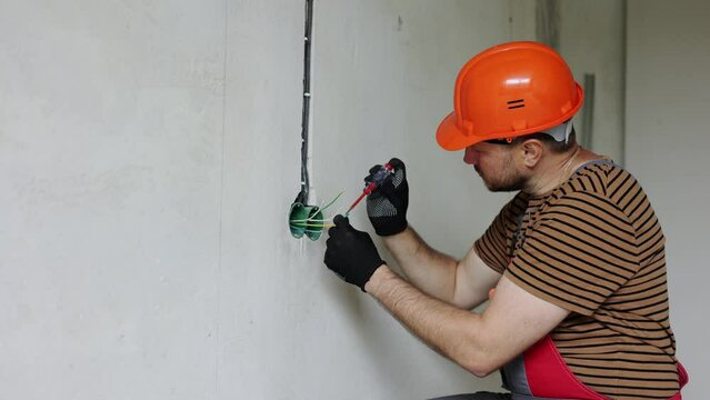 Electrician male in uniform, protective gloves and helmet checks presence of electrical voltage in socket phase uses electrical tester screwdriver.Examining wires in outlet by voltage detector indoor.