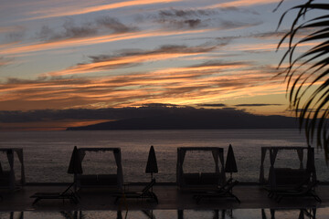Stunning sunset over the Island of La Gomera viewed from Los Gigantes on Tenerife, with sun loungers and palm tress in view.