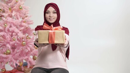 Portrait of caucasian woman in hijab giving wrapped present looking at camera near Christmas tree. Muslim lady standing next to decorated New Year pink fir holds gift box in hands on white background