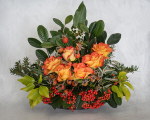 Winter festive floral arrangement. Flame colour roses, berries, greenery and leaves. On pale background. - 690616297