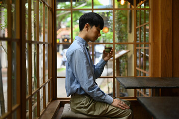A young Asian man sips a small cup of hot tea in a quiet, traditional Japanese-style restaurant or...