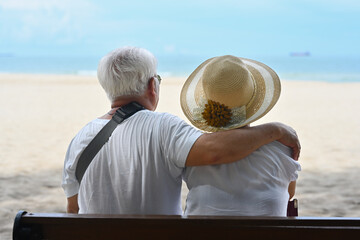Rear view with an Old Asian man hugging his wife while sitting on the beach bench and looking at...