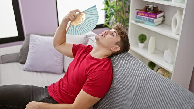 Hot weather woes, young hispanic man suffers, fanning himself with handfan sitting on sofa at home, exposing the struggles of indoor heat