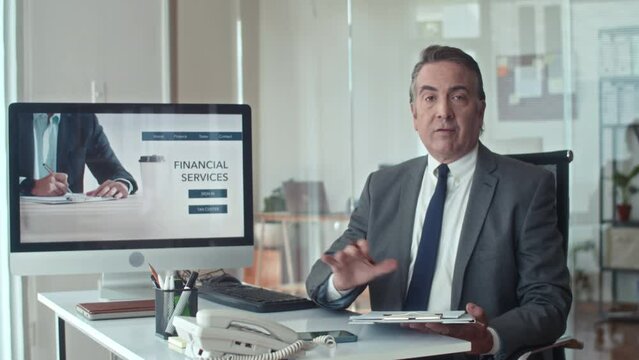 Medium shot of aged entrepreneur pointing at screen of computer with landing page of financial services and talking on camera