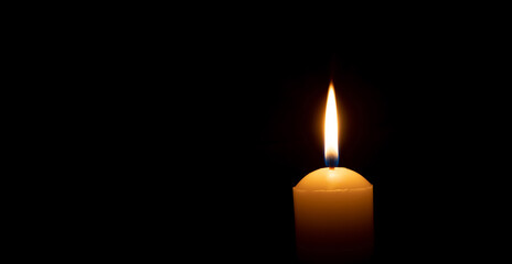 Single burning candle flame or light glowing on a big yellow candle on black or dark background on table in church for Christmas, funeral or memorial service with copy space