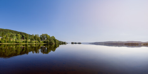 Beautiful panoramic view of the densely forested shores of Lake Snasavatnet in Steinkjer, Norway, reflecting in the lake's tranquil waters under a clear blue summer sky