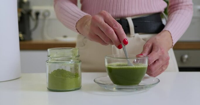 Woman is mixing matcha powder with hot water in glass cup by the spoon