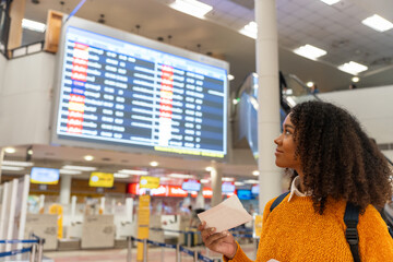 Black woman looks at the flight schedule on a digital monitor in an airport to check the gate and time to board the plane to travel or study abroad. - obrazy, fototapety, plakaty