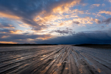 Traces of car tires on a muddy beach, leading to the horizon at nightfall, under a beautifully...
