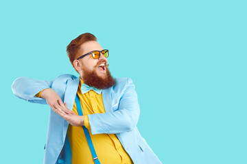 Funny confident fat man in trendy modern outfit having fun at party. Happy overweight bearded guy in cool spectacles, blue suit, yellow shirt, and suspenders dancing on turquoise copy space background - obrazy, fototapety, plakaty
