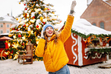 Happy woman in a bright yellow jacket and red lips walks through the Christmas market. Curly-haired tourist woman enjoying winter holidays outdoors. Light all around. Concept of holiday, fun.