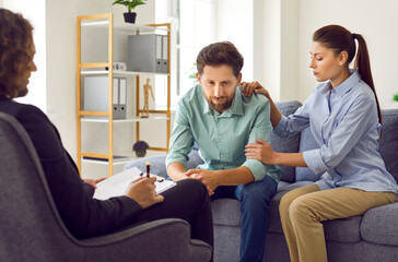 Young married couple sitting on sofa and talking with psychologist doctor. Woman trying to support her husband at counselor's office. Family therapy, relationship and mental health concept.