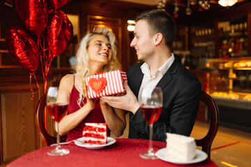 A young couple in love exchanges gifts for Valentine's Day in a cafe at the table. Handsome man and woman on a date in a restaurant. Concept of holiday, love, surprise.