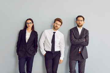 Team of three business people in formal style clothes standing by the office wall. Group portrait of young men and woman in suits, jackets, shirts and ties leaning on a grey wall and looking at camera
