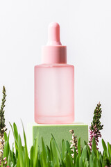 Pink cosmetic bottle with dropper on white background with fresh spring grass and flowers. Natural organic cosmetic concept.