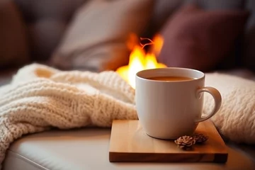 Fotobehang A mug of hot tea stands on a chair with a woolen blanket in a cozy living room with a fireplace. Cozy winter day © PhotoFlex