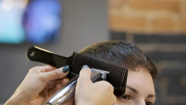 Adjusting the length of short black hair on the side of a woman's head using a black comb and clipper. Careful work with the tool to create a model image in a professional hairdressing salon