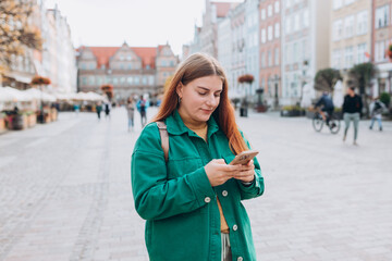 Fototapeta premium Happy cheerful young woman with backpack walking on city street checks her smartphone. Portrait of beautiful 30s girl using smartphone outdoors. Urban lifestyle concept.