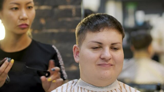 An asian hairdresser carefully arranges black hair with a comb and shaves behind the ear of a fat woman with short hair. Hair care concept in a professional beauty salon and creating a design in image