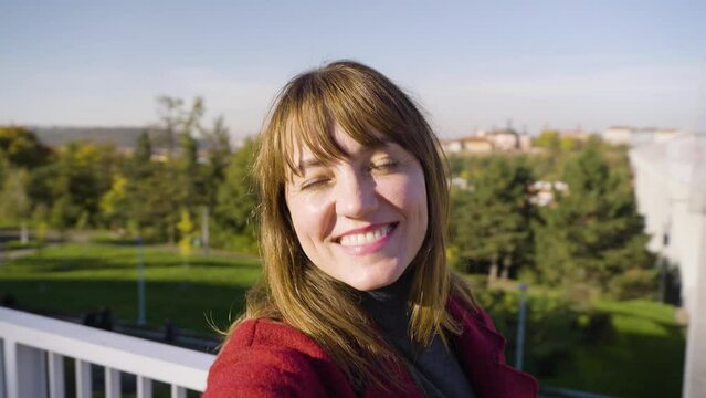 An attractive middle-aged Caucasian woman records herself with a camera and smiles at it - trees and a distant cityscape in the background - selfie shot