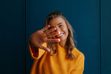 Shy young blond hair woman in yellow sweater stretching out hand on blue background