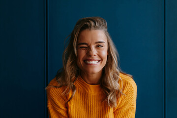 Playful young blond hair woman in yellow sweater winking while having fun on blue wall