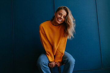 Young blond hair woman in yellow sweater smiling at camera while sitting on blue background